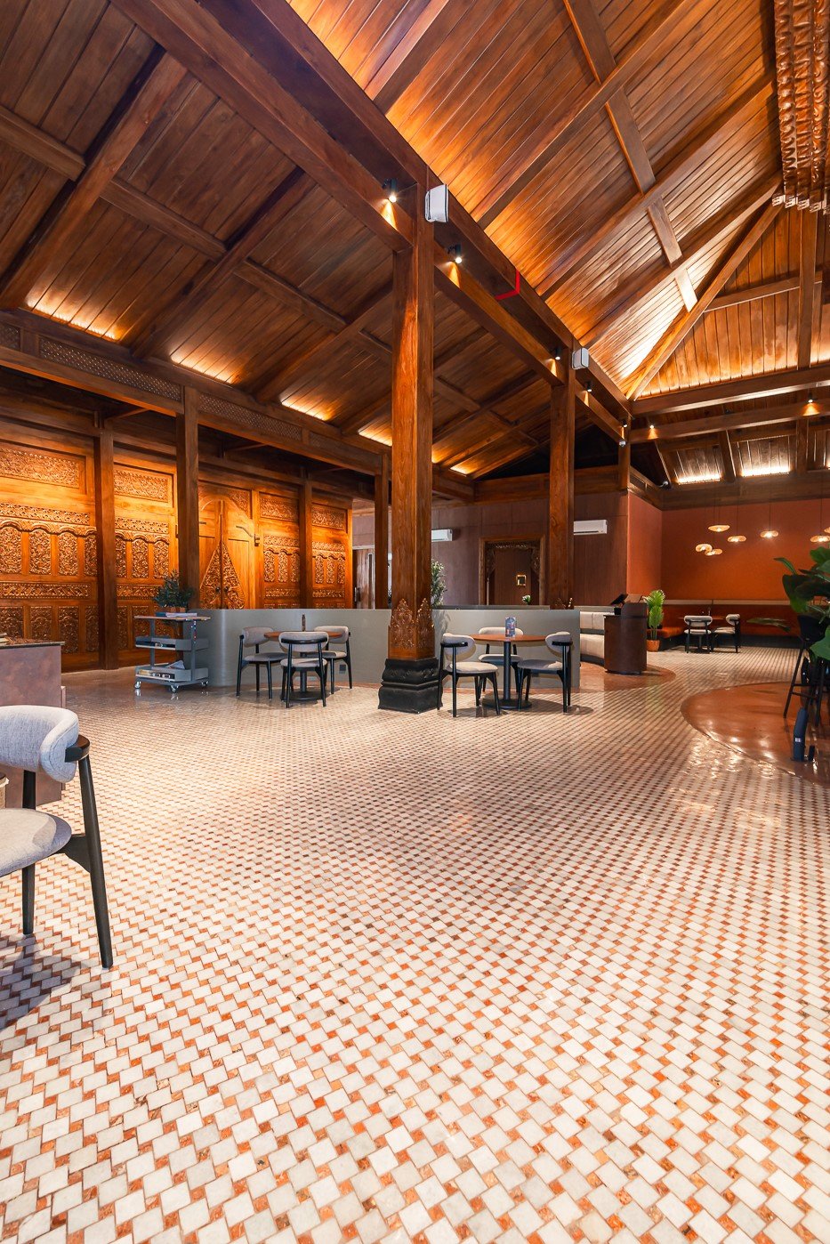Spacious wooden interior with high ceilings, carved panels, and seating, featuring intricate tile flooring.