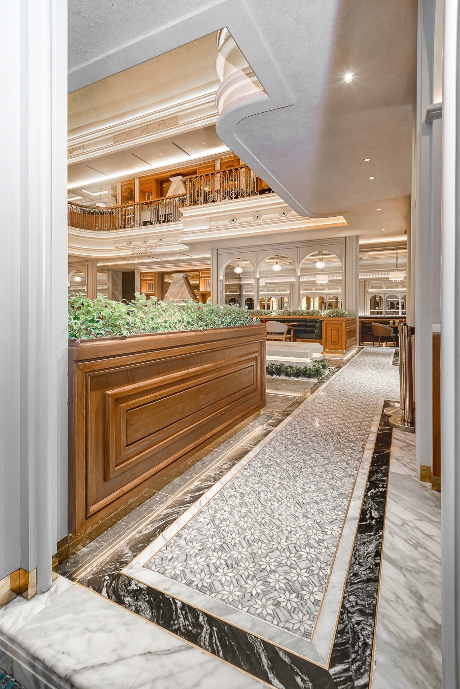 Luxurious hotel lobby with elegant marble flooring and decorative wood paneling, featuring ornate railings and lush greenery.