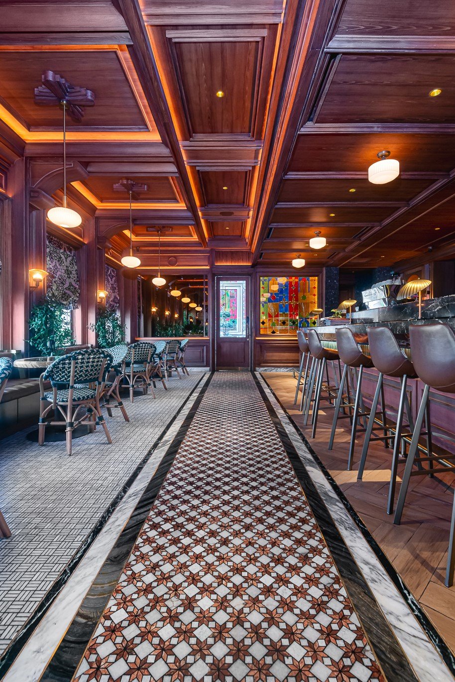 Elegant bar interior with wooden decor, patterned floor, stylish chairs, and vibrant stained glass window.